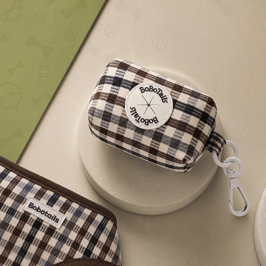 A small BoboTails zip pouch with a brown, beige, and black plaid pattern, featuring a white circular logo patch and a white carabiner clip, displayed on a beige surface.