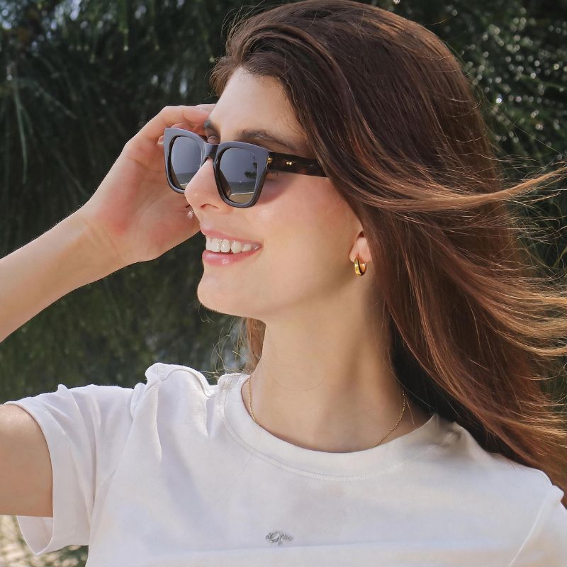 A woman outdoors wearing BOBOTAILS leopard dark sunglasses with a square frame and a white t-shirt, smiling in a sunny setting with green foliage in the background.