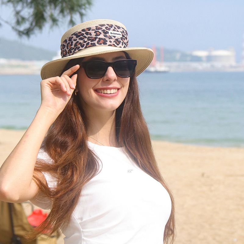 A woman on a beach wearing a beige wide-brim sun hat with a Bobotails leopard-print band, black sunglasses, and a white top, with the ocean and sandy shore in the background.