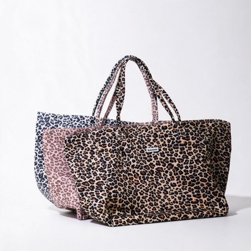 A large tote bag with a leopard print pattern in brown, beige, and black, featuring dual shoulder straps and a small white brand label on the front, displayed against a clean white background.