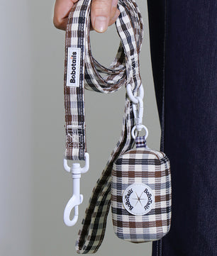 A close-up of a person holding a plaid dog leash and matching small fabric poop bag pouch. The leash and pouch have a brown, beige, and white plaid pattern, with white metal hardware and visible Bobtailis branding on both the strap tag and the pouch patch.