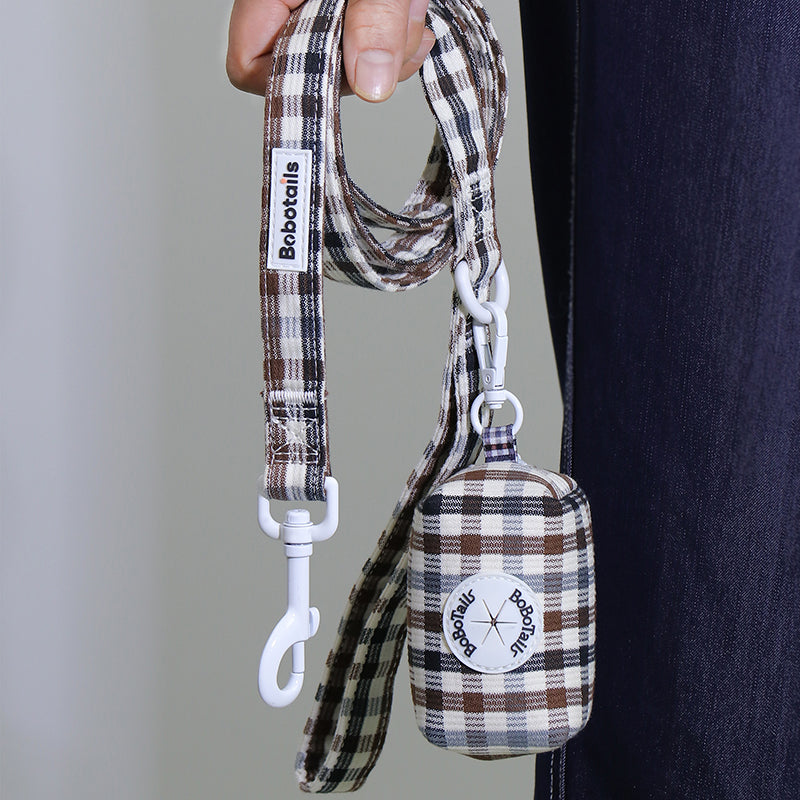 A close-up of a person holding a plaid dog leash and matching small fabric poop bag pouch. The leash and pouch have a brown, beige, and white plaid pattern, with white metal hardware and visible Bobtailis branding on both the strap tag and the pouch patch.