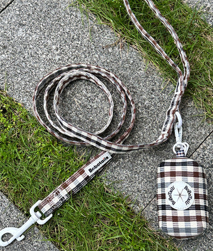 A plaid dog leash with a matching fabric pouch lies on a gray stone path with green grass. The leash has a brown, white, and gray tartan pattern with white metal hardware, and the pouch is labeled 'Bokutou'.