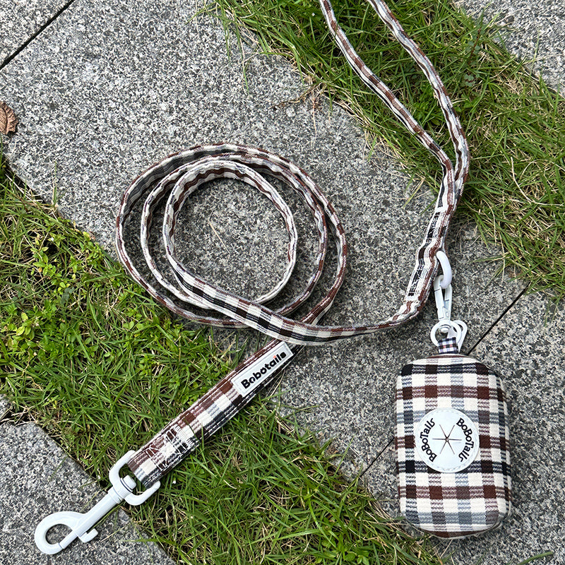 A plaid dog leash with a matching fabric pouch lies on a gray stone path with green grass. The leash has a brown, white, and gray tartan pattern with white metal hardware, and the pouch is labeled 'Bokutou'.