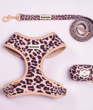 A pet gear set laid out on a light pink background, including a leopard-print harness, matching leash, and small matching pouch. The harness and leash have a beige base with black and brown leopard spots, and the brand name BoboTails is visible on the labels.