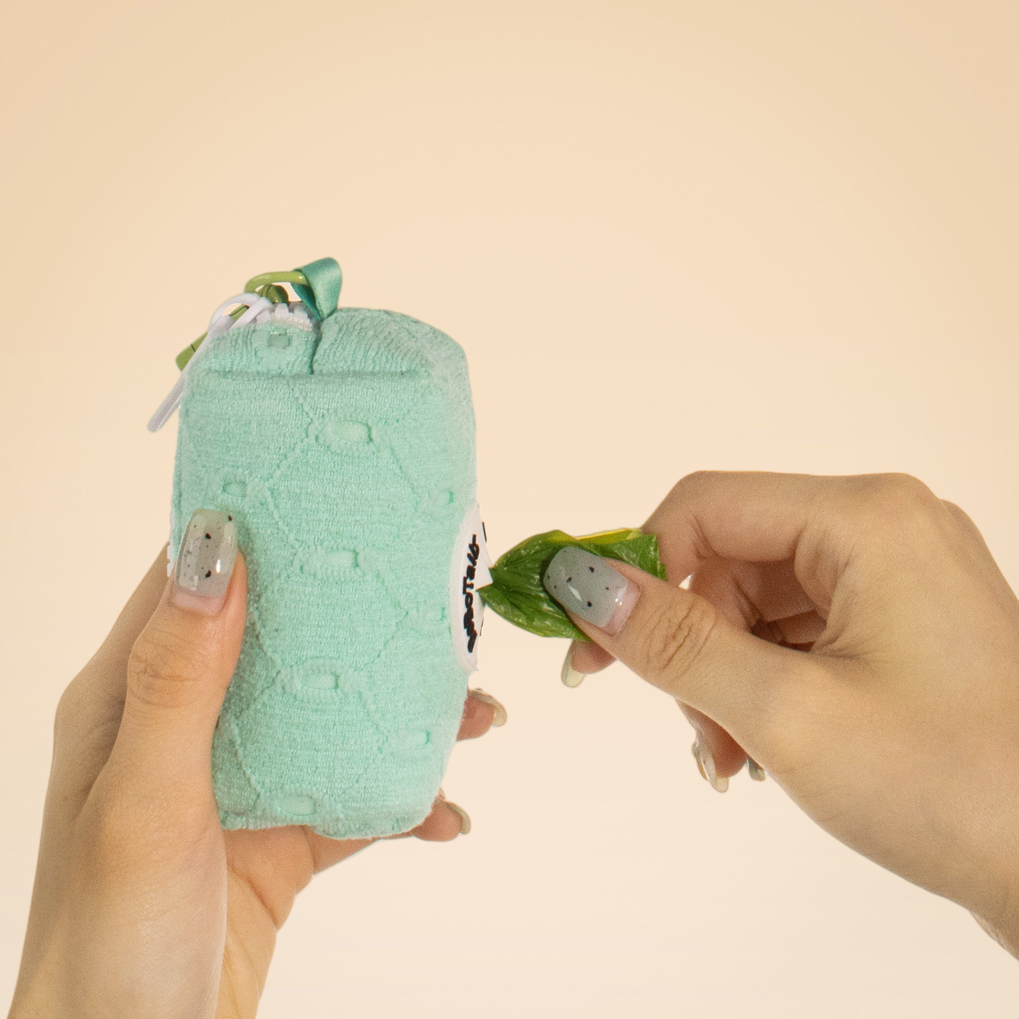 Hand holding a green textured pouch with a small plant-like creature and another hand holding a leaf against a beige background.
