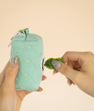 Hand holding a green textured pouch with a small plant-like creature and another hand holding a leaf against a beige background.