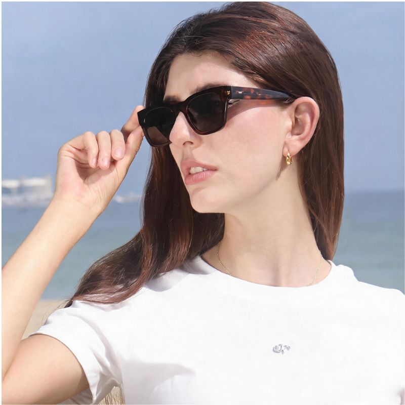 A woman wearing Bobotails leopard tortoiseshell sunglasses and a white t-shirt, posing outdoors with a blurred beach and sky background.