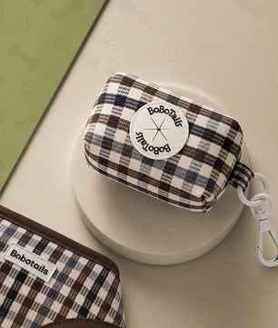 A small BoboTails zip pouch with a brown, beige, and black plaid pattern, featuring a white circular logo patch and a white carabiner clip, displayed on a beige surface.