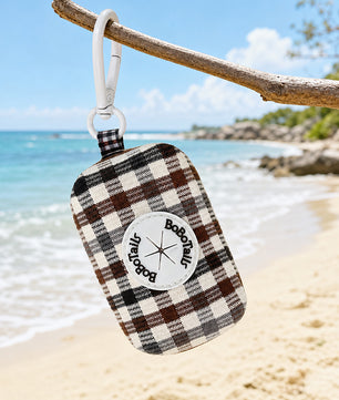 A plaid poop  bag holder with a white clip hangs from a tree branch over a sandy beach with the ocean in the background. The bag has a brown, black, and cream plaid pattern with a white circular patch that reads 'Bobotails.