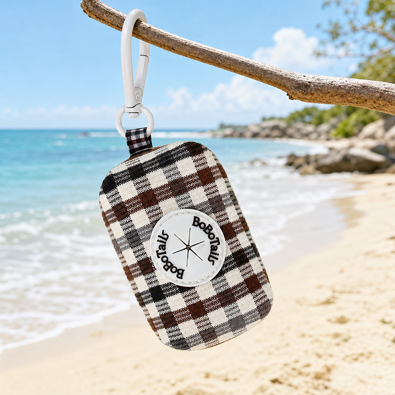 A plaid poop  bag holder with a white clip hangs from a tree branch over a sandy beach with the ocean in the background. The bag has a brown, black, and cream plaid pattern with a white circular patch that reads 'Bobotails.