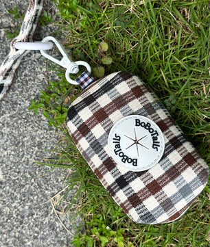 A small plaid pouch with a white circular Boboatou logo patch, attached to a white and gray strap, lying on grass and concrete.