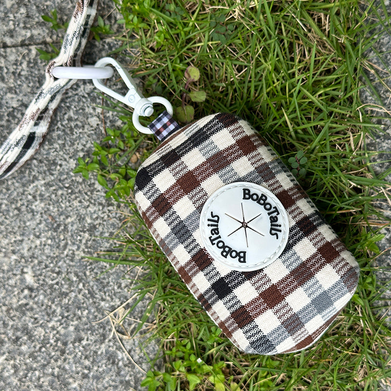 A small plaid pouch with a white circular Boboatou logo patch, attached to a white and gray strap, lying on grass and concrete.