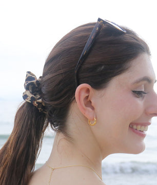 A woman with her hair in a low ponytail wearing a leopard-print scrunchie, photographed outdoors with a blurred beach background. She is also wearing tortoiseshell sunglasses and a small gold hoop earring.