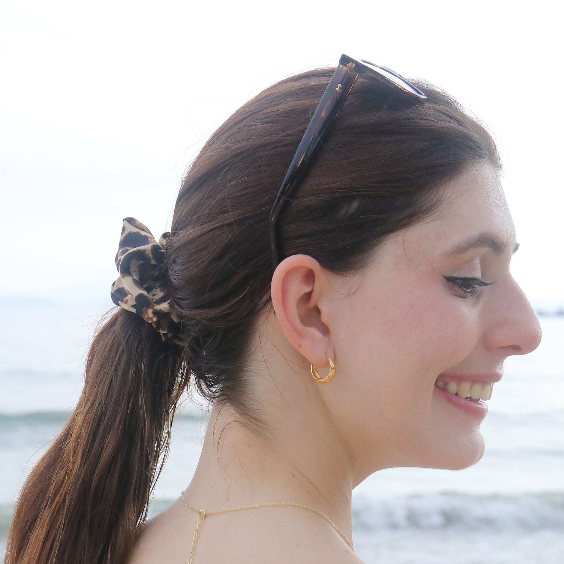 A woman with her hair in a low ponytail wearing a leopard-print scrunchie, photographed outdoors with a blurred beach background. She is also wearing tortoiseshell sunglasses and a small gold hoop earring.