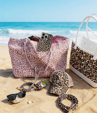 Leopard print accessories including a bag, hat, sunglasses, and bracelet on a sandy beach with ocean view.