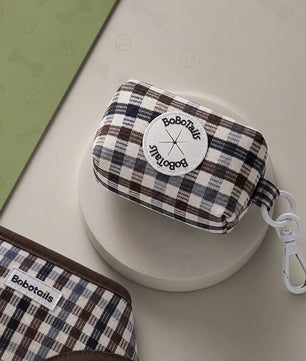 A small BoboTails fabric zip pouch with a brown, cream, and navy plaid pattern, displayed on a round white pedestal. The pouch features a white circular patch with the BoboTails logo and a white plastic carabiner clip attached to the side.