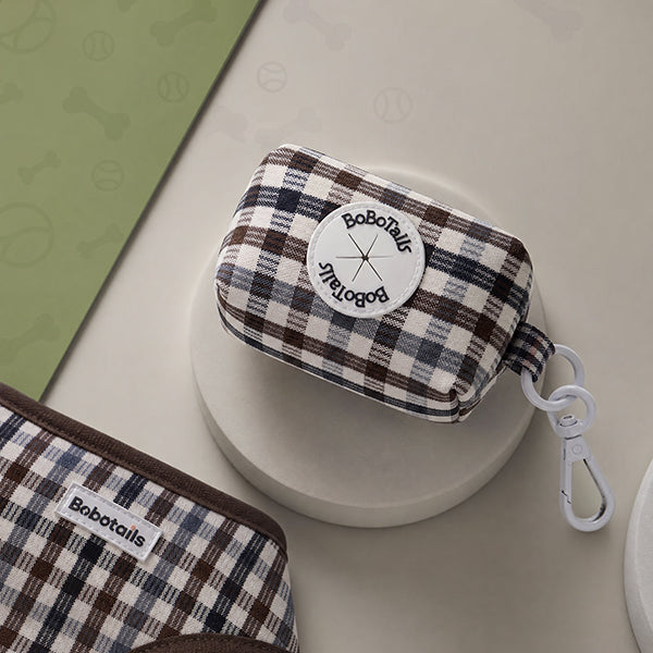 A small BoboTails fabric zip pouch with a brown, cream, and navy plaid pattern, displayed on a round white pedestal. The pouch features a white circular patch with the BoboTails logo and a white plastic carabiner clip attached to the side.