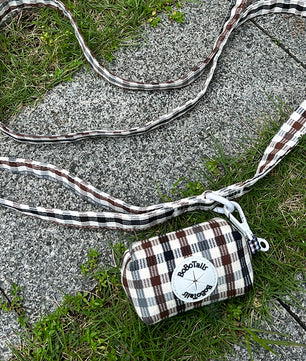 A small plaid dog poop bag holder with a matching long strap lies on gray concrete pavement with green grass around it. The bag has a brown, white, and black plaid pattern with a white circular patch that reads 'Socotally Vintage'.