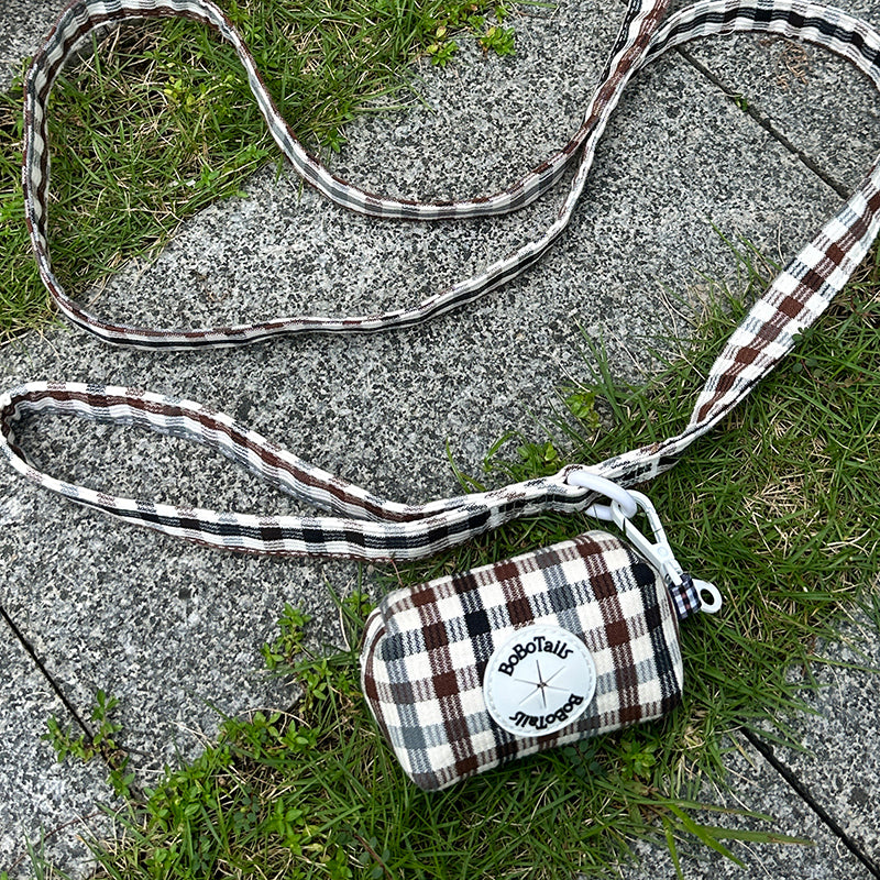 A small plaid dog poop bag holder with a matching long strap lies on gray concrete pavement with green grass around it. The bag has a brown, white, and black plaid pattern with a white circular patch that reads 'Socotally Vintage'.